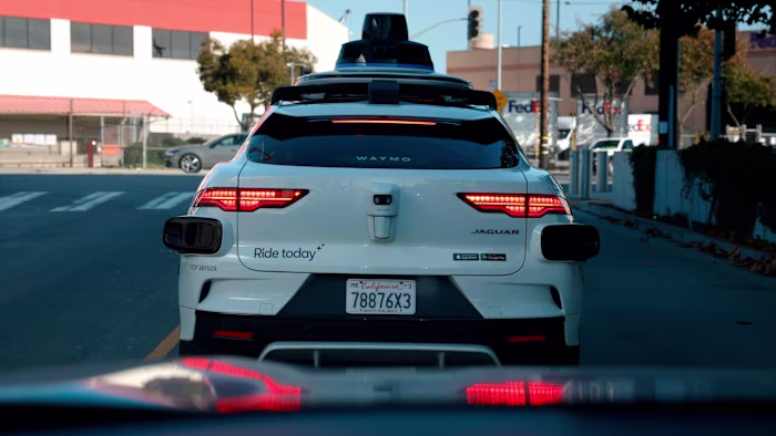 A Waymo self-driving Jaguar robotaxi is stopped at a traffic light, viewed from behind on a city street.