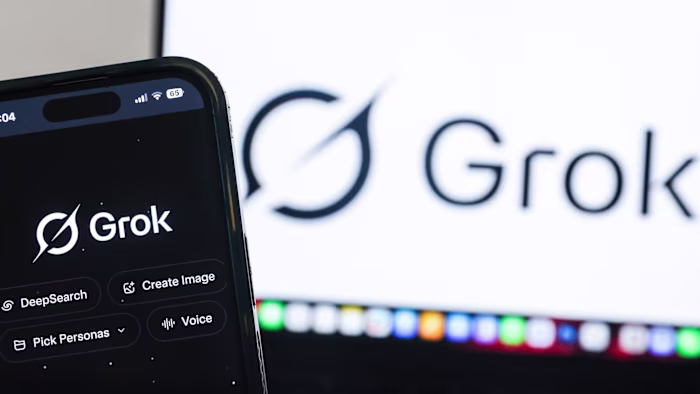 The Grok AI website is shown on a smartphone screen with the Grok logo also displayed on a computer monitor in the background.