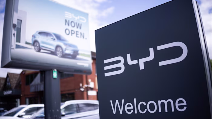 A BYD showroom sign in the foreground with a billboard showing a BYD car and the words "Now open" in the background.