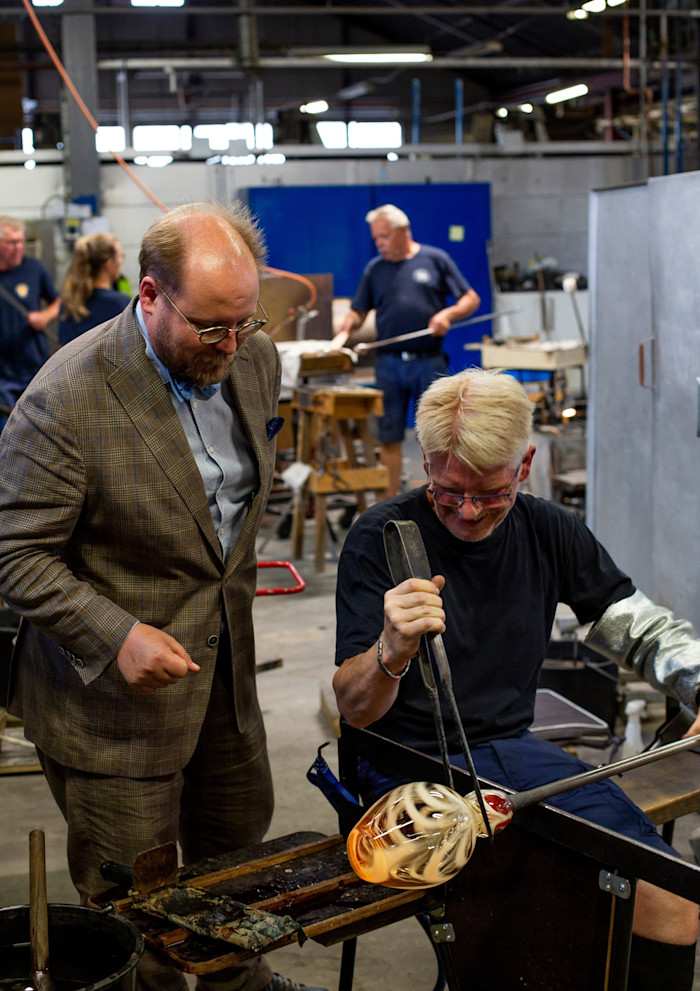 Un soffiatore di vetro modella il vetro fuso con delle pinze mentre un altro uomo osserva da vicino in un laboratorio.