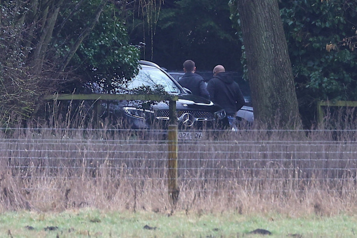 Due uomini, visti di spalle, si allontanano da un'auto nera parcheggiata davanti a una casa, parzialmente oscurata da alberi e recinzioni.