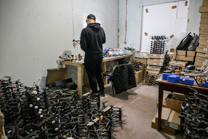 Un tecnico in abiti scuri lavora a un tavolo disordinato in un laboratorio pieno di numerosi droni e scatole di cartone impilate