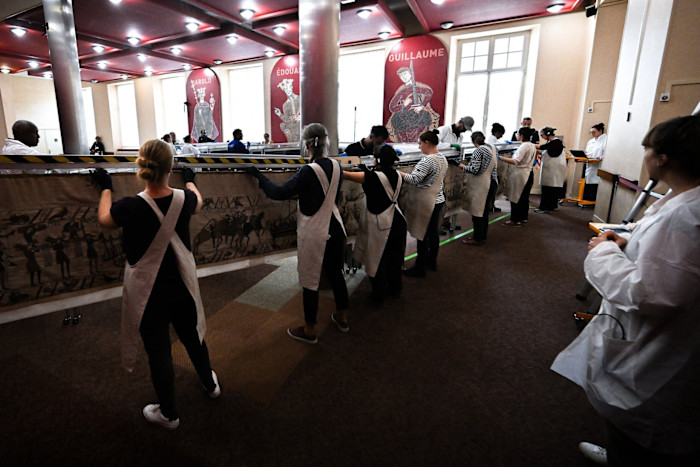 Lavoratori e volontari che indossano grembiuli maneggiano con cura l'Arazzo di Bayeux in preparazione per l'imballaggio all'interno della sala del museo